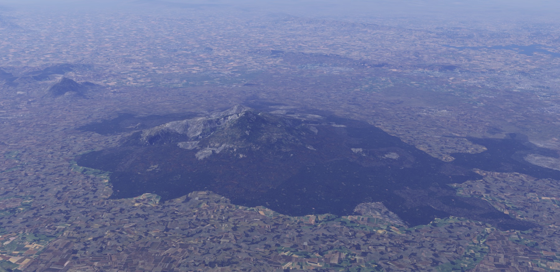 Un volcán aislado de laderas boscosas oscuras, rodeado por completo de campos de cultivo que llegan hasta el borde del bosque; al fondo, una mancha urbana clara y un embalse.